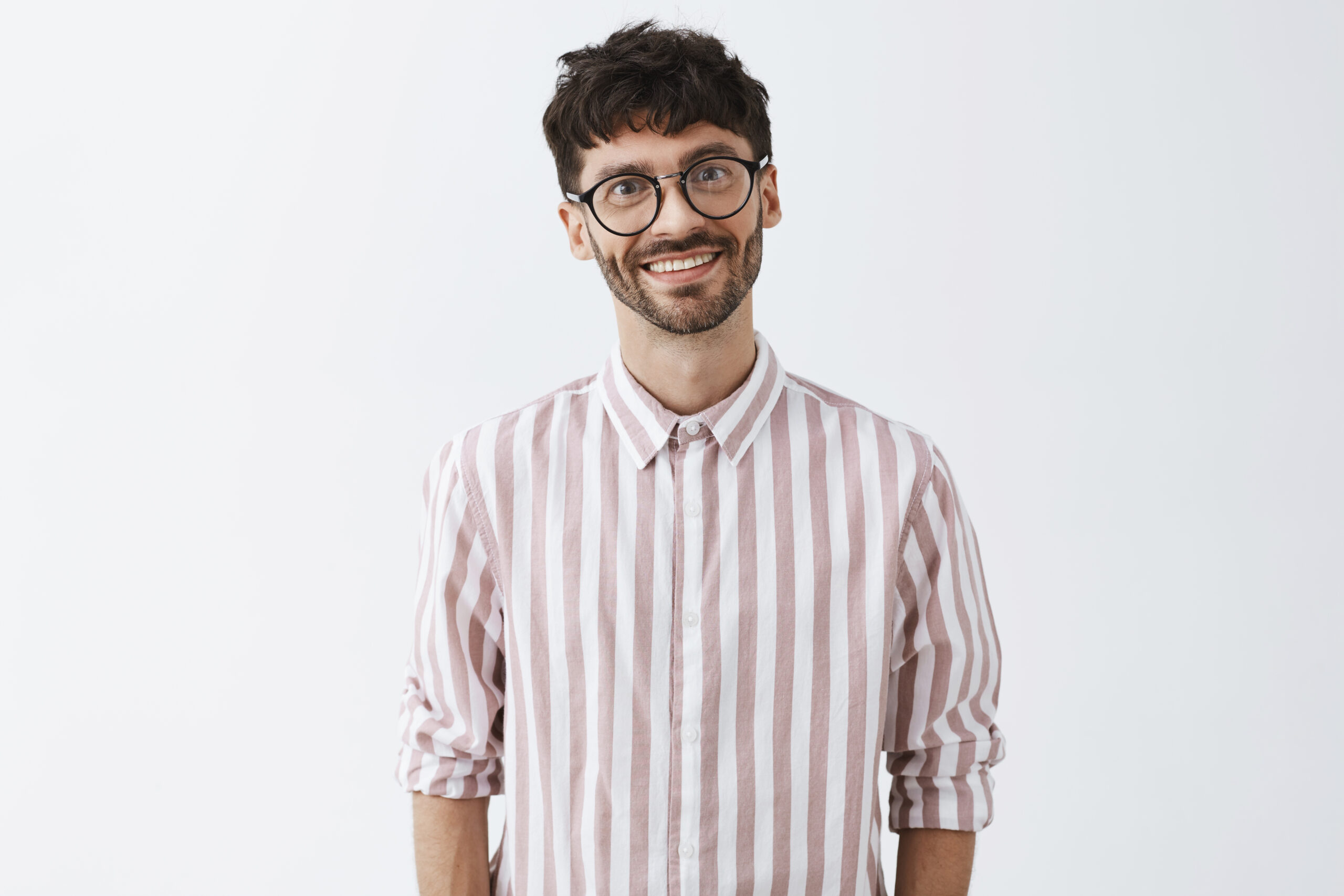 emotive and funny cute bearded guy with dark stylish hairstyle tilting head and smiling with polite and warm expression being in love with girl listening with affection talk over gray wall
